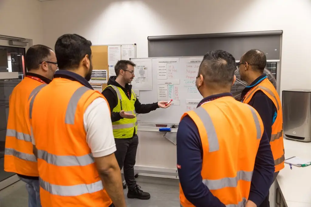 Outdoor workers in high-visibility vests gathered around a whiteboard during a team briefing.