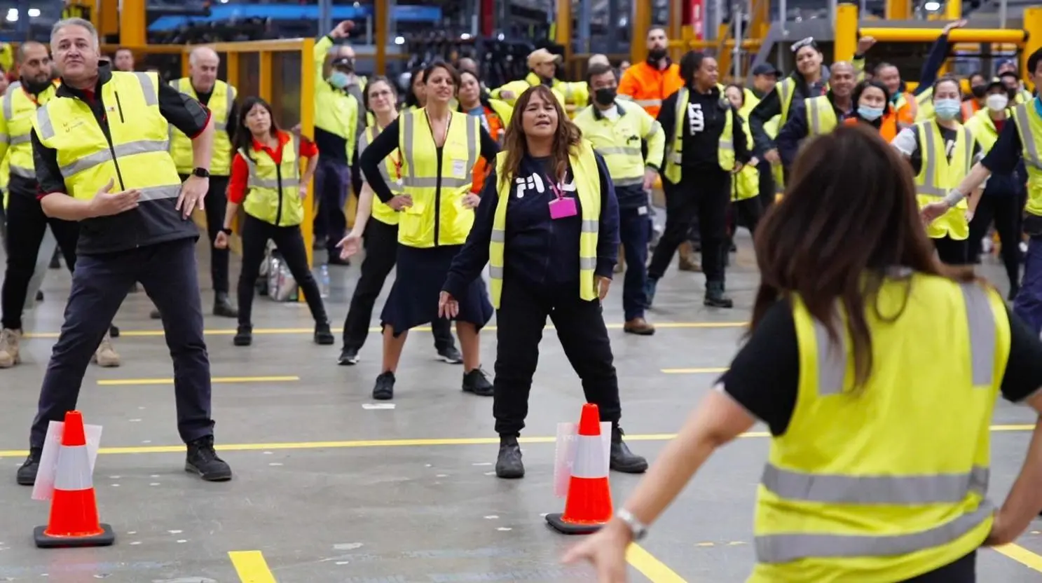A team leader running a shift-start warm-up for warehouse workers in high-vis vests