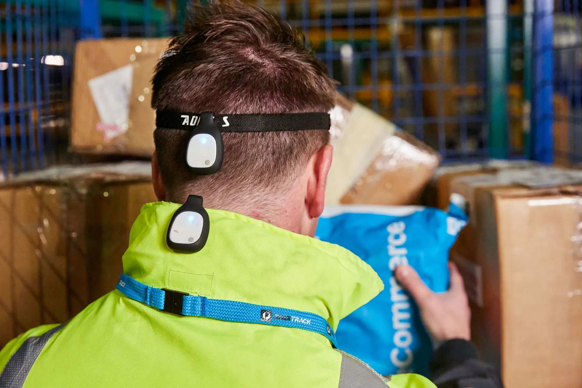 A warehouse worker wearing EMP motion sensors on the head and upper back while lifting a carton.