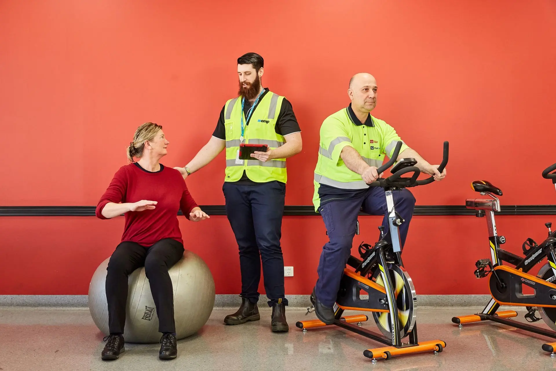 An EMP Onsite clinician guiding two workers through a conditioning session on exercise bikes and a stability ball.